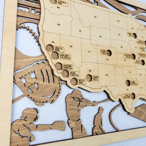 May include: A wooden map of the United States with baseball stadiums marked with holes. The map is decorated with a baseball glove and two baseball players. The text on the map includes the names of the stadiums, such as "Dodger Stadium", "Angel Stadium", and "Petco Park".