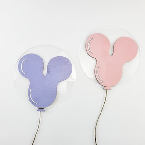 K&ouml;nnte beinhalten: Zwei Holz-Ballon-Dekorationen, eine lila und eine rosa, in Mickey-Maus-Form, die an klaren Acrylkreisen h&auml;ngen.
