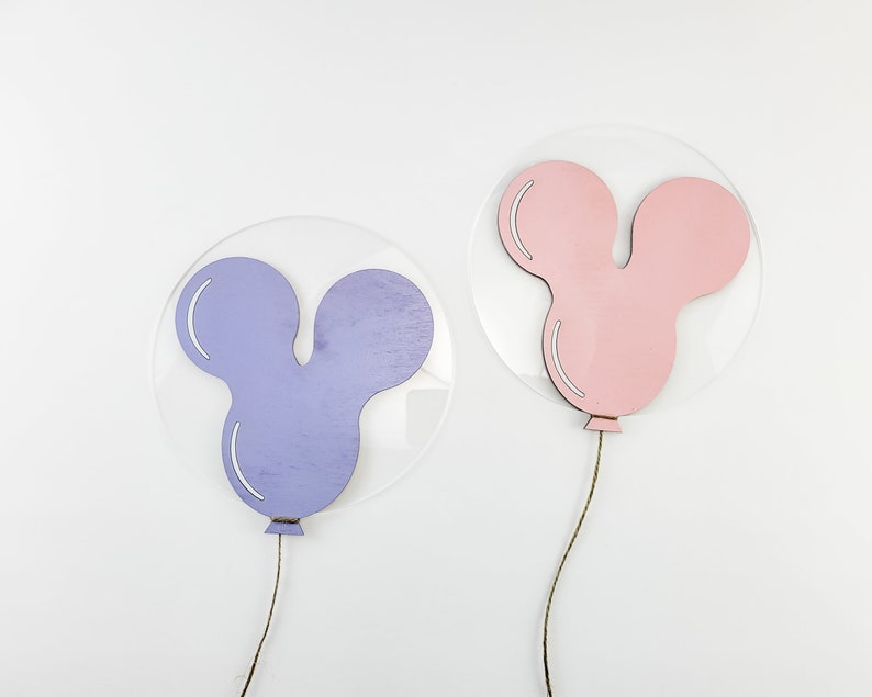 K&ouml;nnte beinhalten: Zwei Holz-Ballon-Dekorationen, eine lila und eine rosa, in Mickey-Maus-Form, die an klaren Acrylkreisen h&auml;ngen.