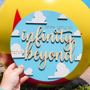 May include: A round, light blue wooden sign with the phrase "I love you to infinity and beyond" in a whimsical font. White cloud cutouts are scattered across the sign, which is held in front of a large yellow ball.