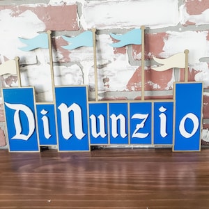 May include: A wooden sign in the shape of a castle with the name "DiNunzio" in white, stylized lettering. The castle is blue with beige accents and small flags. The sign is displayed against a brick wall.