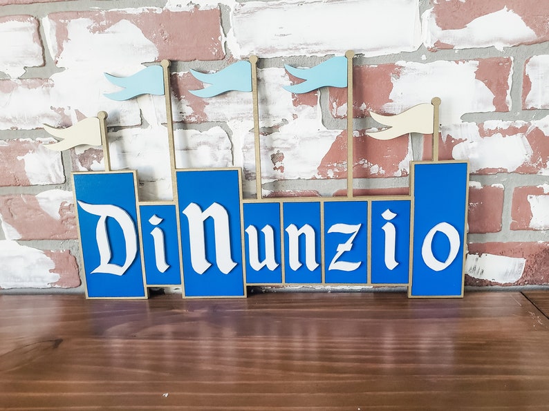 May include: A wooden sign in the shape of a castle with the name "DiNunzio" in white, stylized lettering. The castle is blue with beige accents and small flags. The sign is displayed against a brick wall.