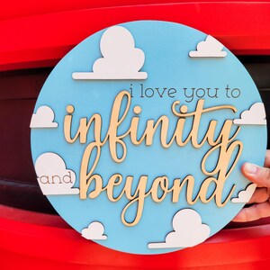May include: A round wooden sign with a light blue background and white cloud cutouts. The sign features the phrase "i love you to infinity and beyond" in a mix of black and natural wood tones.