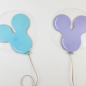 K&ouml;nnte beinhalten: Zwei ausgeschnittene Holzballons, einer hellblau und einer lila, mit befestigten Schn&uuml;ren. Die Ballons sind in Form von Mickey-Maus-Ohren.