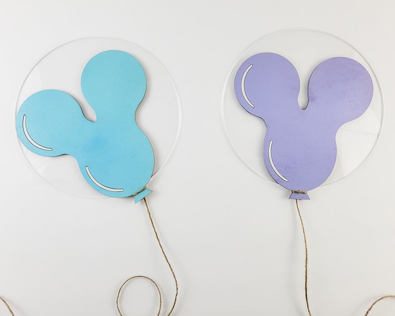K&ouml;nnte beinhalten: Zwei ausgeschnittene Holzballons, einer hellblau und einer lila, mit befestigten Schn&uuml;ren. Die Ballons sind in Form von Mickey-Maus-Ohren.