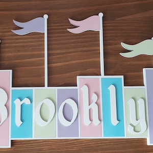 May include: A pink, blue, green, and purple castle-shaped name sign with the name "Brooklyn" spelled out in white letters.