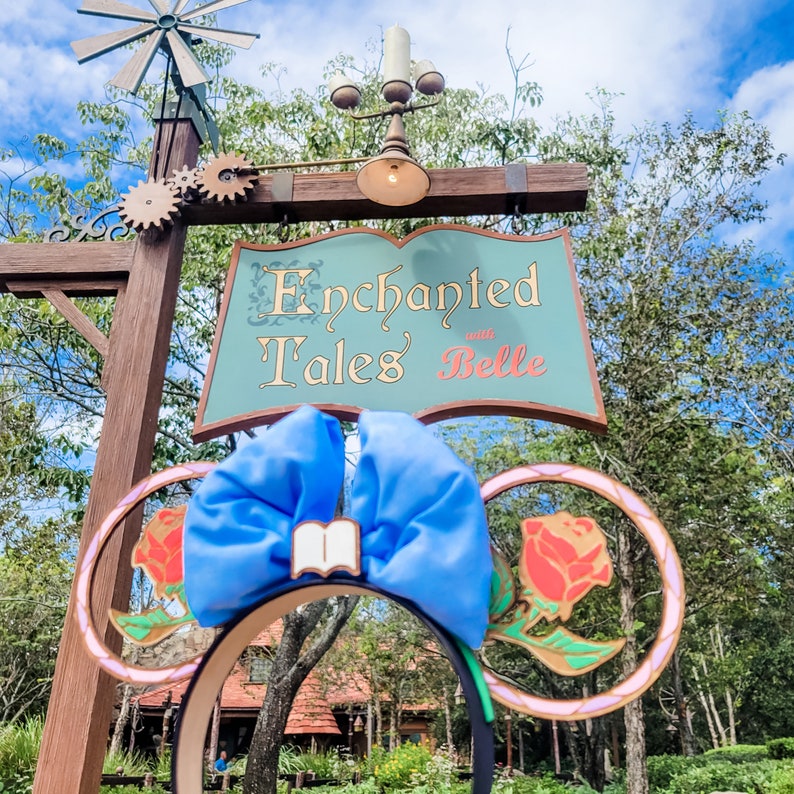 May include: A pair of blue and pink Minnie Mouse ears with a book charm and a red rose design. The ears are in front of a sign that reads "Enchanted Tales with Belle".