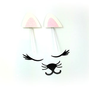 May include: A set of white felt cat ears with pink felt inside, black felt eyelashes, and a black felt nose. The ears are on white sticks.