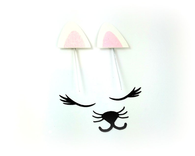 May include: A set of white felt cat ears with pink felt inside, black felt eyelashes, and a black felt nose. The ears are on white sticks.