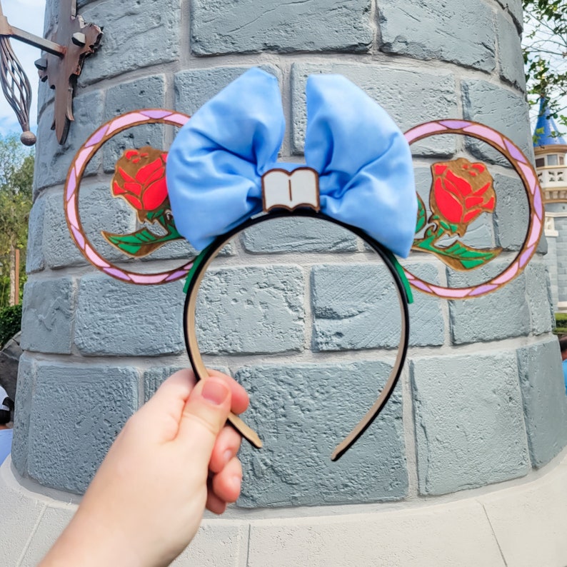 May include: Light blue bow headband with a book charm and two rose-shaped charms with stained glass designs. The headband is made of a soft, flexible material and is perfect for adding a touch of whimsy to any outfit.