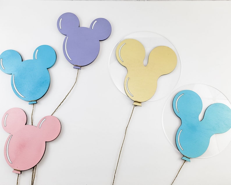 K&ouml;nnte beinhalten: F&uuml;nf Mickey-Maus-f&ouml;rmige Ballons aus Holz in verschiedenen Farben. Die Ballons h&auml;ngen an Schn&uuml;ren. Die Ballons sind hellblau, lila, gelb und zwei sind blau.