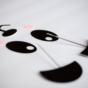 May include: Black felt panda face craft pieces with pink felt circles and white felt circles. The pieces are attached to white skewers.