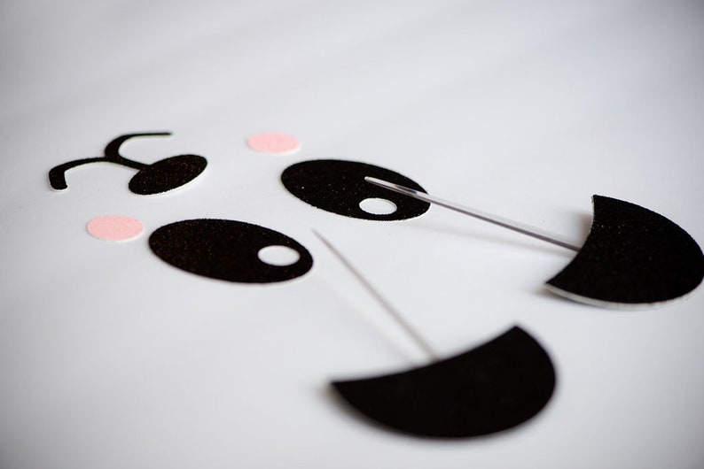 May include: Black felt panda face craft pieces with pink felt circles and white felt circles. The pieces are attached to white skewers.