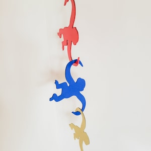 Puede incluir: Tres siluetas de monos de madera colgando de una cuerda. El mono de arriba es rojo, el mono del medio es azul y el mono de abajo es dorado.
