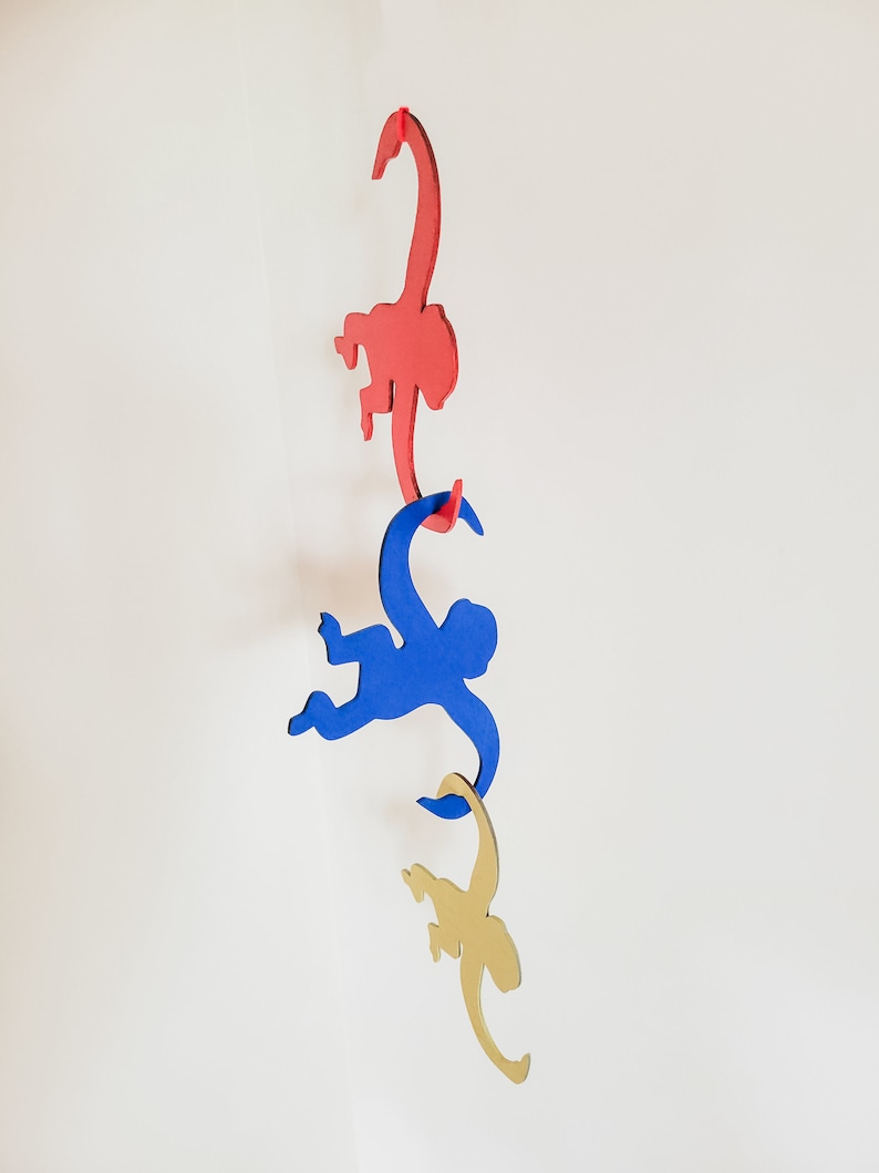 Puede incluir: Tres siluetas de monos de madera colgando de una cuerda. El mono de arriba es rojo, el mono del medio es azul y el mono de abajo es dorado.