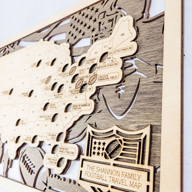 K&ouml;nnte beinhalten: Ein Wandkunstwerk aus Holz mit einer Karte der Vereinigten Staaten und den Standorten von Footballstadien. Die Karte ist lasergeschnitten mit einem mehrschichtigen Design, das die Bundesstaaten und Stadionnamen zeigt. Der Text "THE SHANNON FAMILY FOOTBALL TRAVEL MAP" wird unten angezeigt.