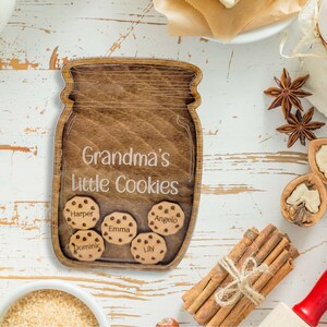 May include: Wooden jar-shaped sign with the text "Grandma's Little Cookies." The sign has five cookie-shaped cutouts with names: Harper, Emma, Angelo, Dominic, and Lily. The sign is brown with a light wood grain.