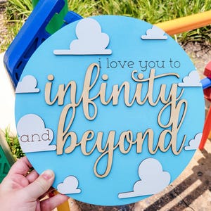 May include: A round, light blue wooden sign with the phrase "i love you to infinity and beyond" in cursive and block letters. White cloud cutouts are placed around the edge of the sign.