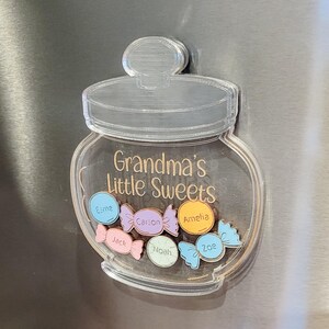 May include: Clear acrylic jar-shaped magnet with the text "Grandma's Little Sweets." The jar contains colorful candy-shaped magnets with names: Esme, Jack, Carson, Noah, Amelia, and Zoe. The magnet is on a silver surface.