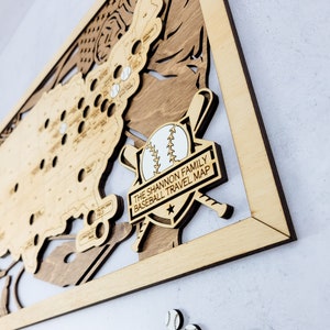 May include: A wooden baseball travel map with a white baseball and crossed bats in the center. The map is framed in wood and has the text "The Shannon Family Baseball Travel Map" on a black banner.
