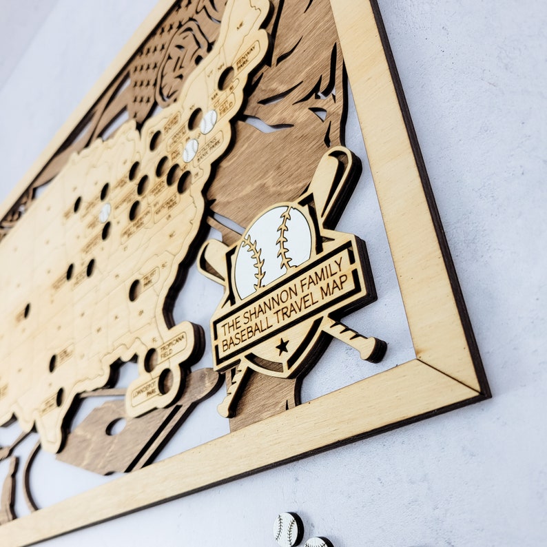 May include: A wooden baseball travel map with a white baseball and crossed bats in the center. The map is framed in wood and has the text "The Shannon Family Baseball Travel Map" on a black banner.