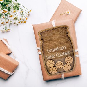 May include: A wooden jar-shaped sign with the text "Grandma's Little Cookies" and cookie-shaped name tags. The sign is placed on a wrapped gift with twine. White daisies are in the background.