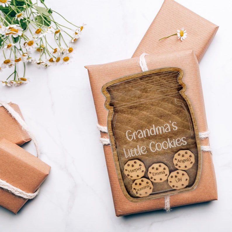 May include: A wooden jar-shaped sign with the text "Grandma's Little Cookies" and cookie-shaped name tags. The sign is placed on a wrapped gift with twine. White daisies are in the background.