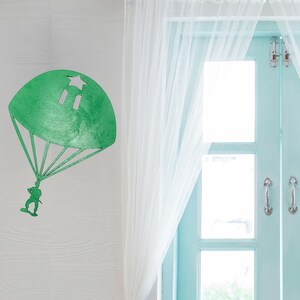 Puede incluir: Decoraci&oacute;n de pared de madera verde que representa un paraca&iacute;das con una estrella y dos rect&aacute;ngulos recortados en la cubierta. Una peque&ntilde;a figura est&aacute; suspendida debajo. La decoraci&oacute;n est&aacute; montada en una pared blanca.