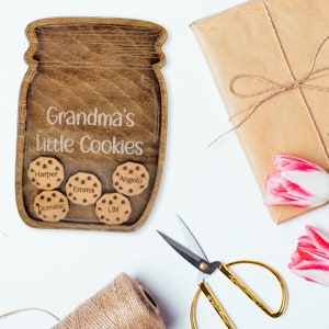 May include: A wooden jar-shaped sign with the text "Grandma's Little Cookies" and the names Harper, Emma, Angelo, Dominic, and Lily on cookie-shaped cutouts. The sign is surrounded by a wrapped gift, tulips, scissors, and twine.