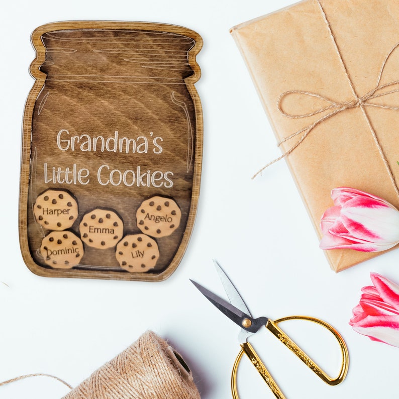 May include: A wooden jar-shaped sign with the text "Grandma's Little Cookies" and the names Harper, Emma, Angelo, Dominic, and Lily on cookie-shaped cutouts. The sign is surrounded by a wrapped gift, tulips, scissors, and twine.