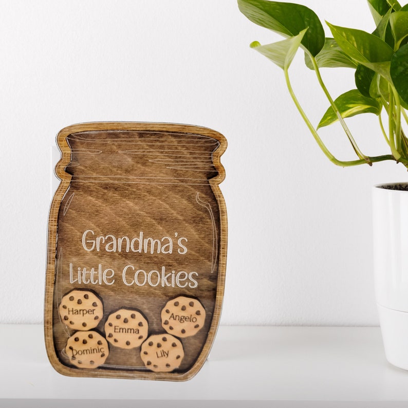 May include: Wooden cookie jar-shaped sign with the words "Grandma's Little Cookies" and five cookie-shaped name tags. The jar is made of dark brown wood, and the names are engraved in a light color. A decorative item.