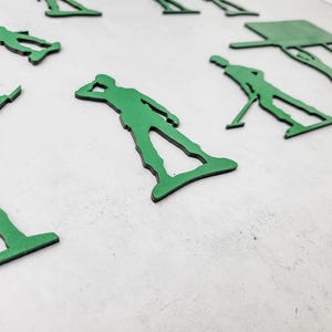 Puede incluir: Recortes de soldados de juguete verdes en varias poses, incluyendo saludar y apuntar. Los recortes est&aacute;n dispuestos sobre una superficie blanca. Los soldados son de un color verde vibrante con bordes negros, creando un contraste distintivo.