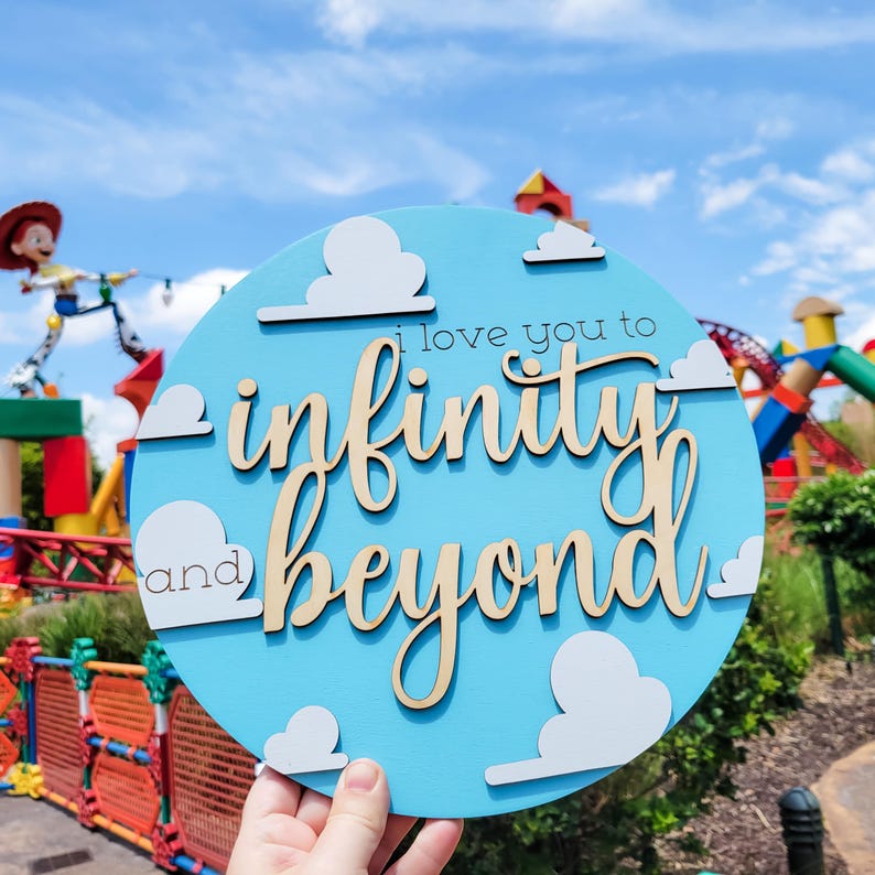 May include: A round, light blue wooden sign with the phrase "I love you to infinity and beyond" in a whimsical font. White cloud cutouts are placed around the text. The sign is held up against a backdrop of a theme park.