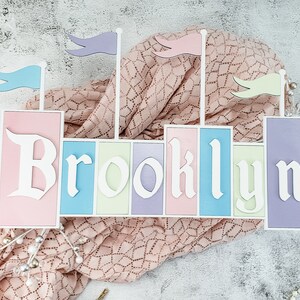 May include: A wooden castle-shaped sign with the name "Brooklyn" spelled out in white letters on pastel-colored panels. The sign is decorated with colorful flags.
