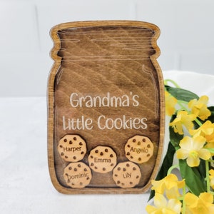 May include: Wooden jar-shaped sign with the text "Grandma's Little Cookies." The sign features five cookie-shaped cutouts with names: Harper, Emma, Angelo, Dominic, and Lily. The sign is made of wood with a dark brown stain.