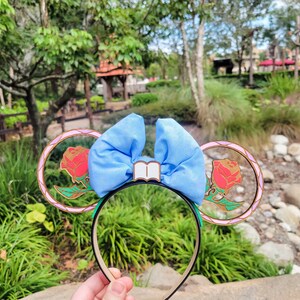 May include: A pair of blue and gold mouse ears with a bow and a book charm. The ears feature a rose design with a stained glass effect.