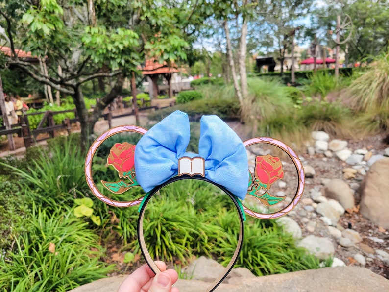 May include: A pair of blue and gold mouse ears with a bow and a book charm. The ears feature a rose design with a stained glass effect.