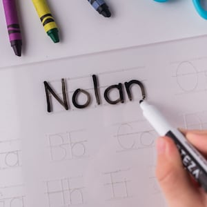 May include: A white dry-erase board with the name "Nolan" written in black marker. The board has a grid of dotted lines for practicing writing letters. A white dry-erase marker is being used to trace over the letters.