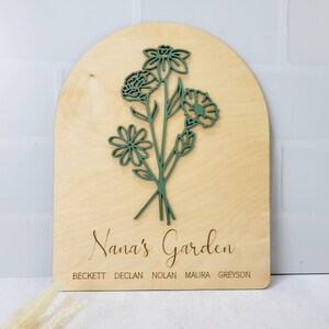 Könnte beinhalten: Ein Holzschild mit einem grünen Blumenmuster und dem Text "Nana's Garden" mit den Namen Beckett, Declan, Nolan, Maura und Greyson darunter.