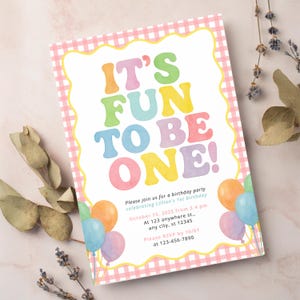 Puede incluir: Una invitación de cumpleaños con el texto "IT'S FUN TO BE ONE!" en letras coloridas y redondeadas. La invitación tiene un borde de cuadros rosa e incluye detalles de la fiesta e ilustraciones de globos.