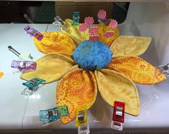 May include: A flower-shaped fabric pincushion with yellow and orange patterned petals, a blue center, and pink tulip-shaped pins. The pincushion is secured with various colorful plastic sewing clips and rests on a white sewing machine surface.