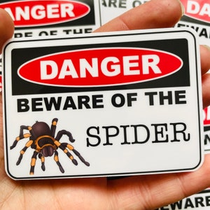 May include: A white warning sign with black and red borders. The sign reads "Danger Beware of the Spider" with a cartoon image of a black and yellow spider.