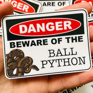 Peut inclure: Autocollant rectangulaire avec un design de panneau d'avertissement. Il présente un ovale rouge avec le mot "DANGER" en blanc, au-dessus du texte "BEWARE OF THE BALL PYTHON". Une illustration de serpent marron et beige est à gauche.