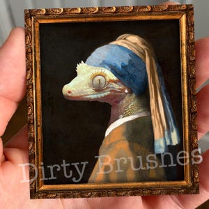 Puede incluir: Una pintura de un gecko con un pañuelo azul y blanco con un pendiente de perla. El gecko está pintado con un estilo realista y se coloca sobre un fondo oscuro. La pintura está enmarcada en un marco dorado.