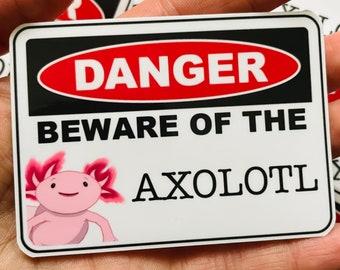Könnte beinhalten: Ein weißer Aufkleber mit einem roten und schwarzen Warnschild, auf dem "GEFAHR ACHTUNG VOR DEM AXOLOTL" steht. Ein Cartoon-Axolotl mit rosa Haut und roten Kiemen befindet sich in der linken unteren Ecke des Aufklebers.