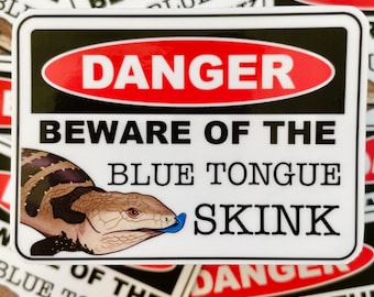 Peut inclure: Un autocollant blanc avec du texte noir et rouge qui dit "Danger Attention au lézard à langue bleue". Une image de dessin animé d'un lézard à langue bleue est représentée sur l'autocollant.