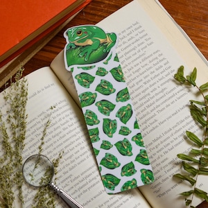 Puede incluir: Un marcapáginas caprichoso con una rana verde posada en una hoja. El marcapáginas tiene un patrón repetido de ranas en hojas. El marcapáginas está colocado en un libro abierto.