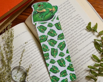 Könnte beinhalten: Ein skurriles Lesezeichen mit einem grünen Frosch, der auf einem Blatt sitzt. Das Lesezeichen hat ein sich wiederholendes Muster von Fröschen auf Blättern. Das Lesezeichen liegt auf einem offenen Buch.
