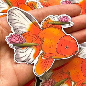 May include: A sticker of a goldfish with white and orange scales swimming in a pond with pink lily pads. The goldfish is facing right and has a large, round body. The sticker is on a hand.