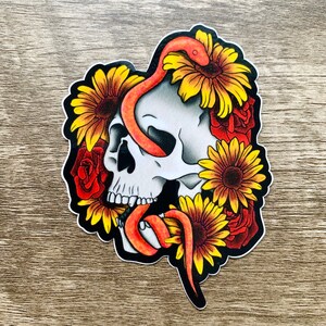Könnte beinhalten: Ein Aufkleber mit einem weißen Schädel, um den sich eine rote Schlange windet. Der Schädel ist von gelben Sonnenblumen und roten Rosen umgeben.